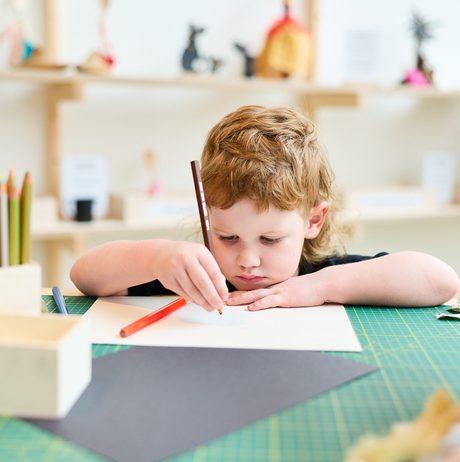 A young child with curly hair sits at a crafting table, focused on drawing with a coloured pencil. Surrounding them are art supplies, including coloured pencils and sheets of paper, in a bright, creative space.