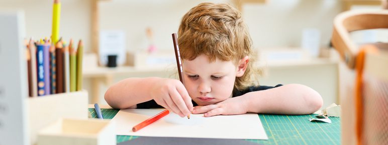 A young child with curly hair sits at a crafting table, focused on drawing with a coloured pencil. Surrounding them are art supplies, including coloured pencils and sheets of paper, in a bright, creative space.