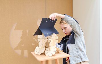 A young child with short hair is focused on assembling a wooden model on a small table. The model features wheels and a roof, and the child is carefully attaching parts with metal clips, showcasing creativity and concentration.