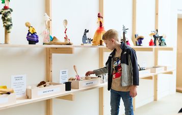 A young boy examines a display of handmade figurines and everyday objects on wooden shelves in a bright, modern exhibition space. The items include colourful dolls and various crafted objects, inviting interaction and exploration.