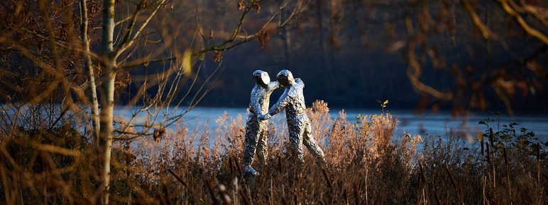 Two silver figures wearing spacesuits in an embrace. Autumnal foliage and a lake are behind the sculpture.