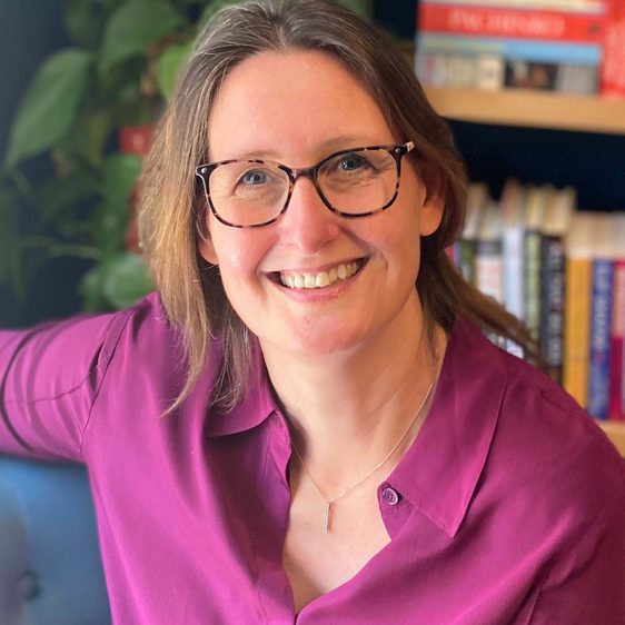 A woman with shoulder length brown hair and glasses, wearing a pink shirt.