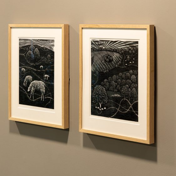 Two framed lino cut prints of landscapes on a grey wall.