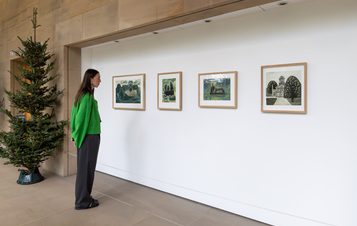 A woman in a green jumper looking at four framed prints on a white wall. A Christmas tree is at the left hand side.