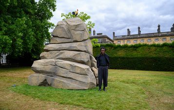 A man stands beside a large, realistic rock sculpture in a grassy area, with trees and a historic building in the background. The sky is overcast, adding a dramatic effect to the scene.