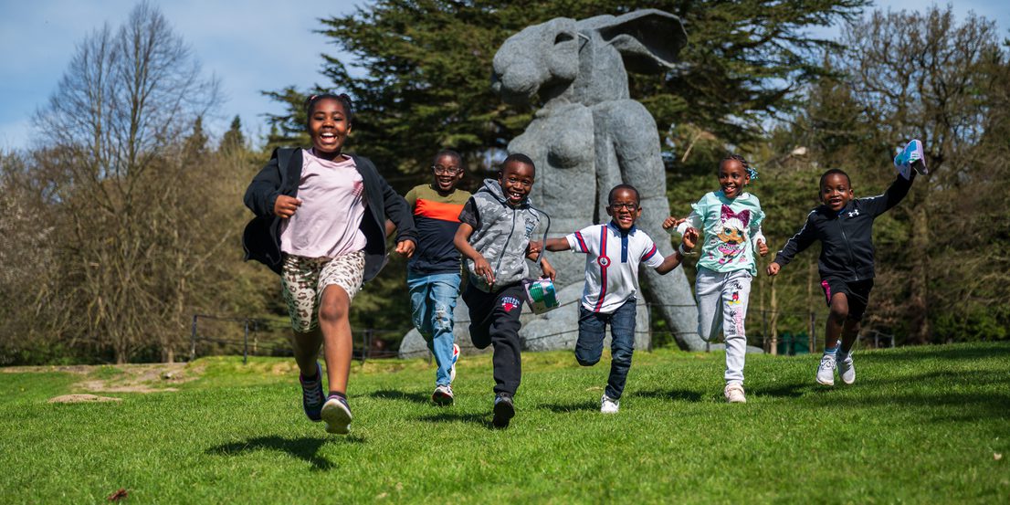 A group of six children joyfully run across a grassy field, smiling and laughing. In the background, a large sculpture of a rabbit stands among trees under a clear blue sky, creating a lively and playful atmosphere.