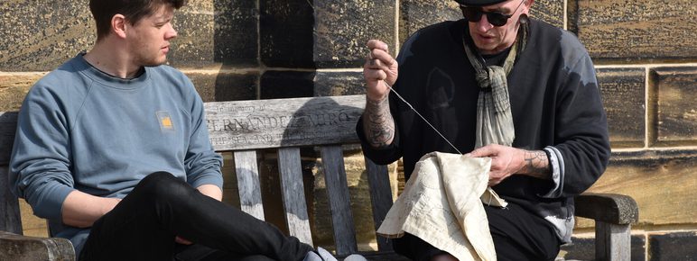 A person in a black beret and scarf sits on a wooden bench, sewing a piece of fabric. Next to him, a person in a grey sweatshirt watches intently. They are outdoors, with a stone wall in the background, enjoying a sunny day.