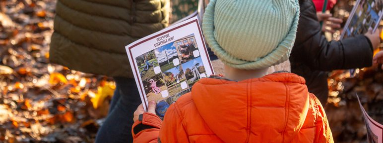 A child wearing a winter coat and wooly hat holding a trail sheet with images of sculptures on it.