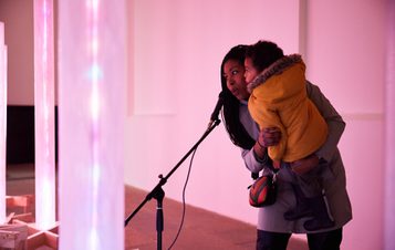 An adult holding a toddler, singing into a microphone surrounded by glowing columns.