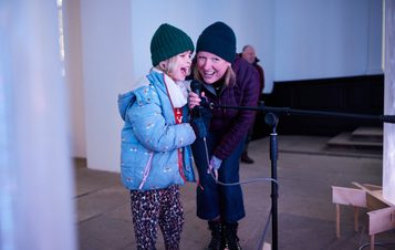 An adult and child singing into a microphone surrounded by glowing columns.