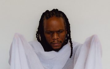 A man with braided hair is holding a sheer, light-coloured fabric in front of him. He appears contemplative, with his eyes closed, and is dressed in a simple, flowing garment. The background is plain and neutral, enhancing the focus on him.