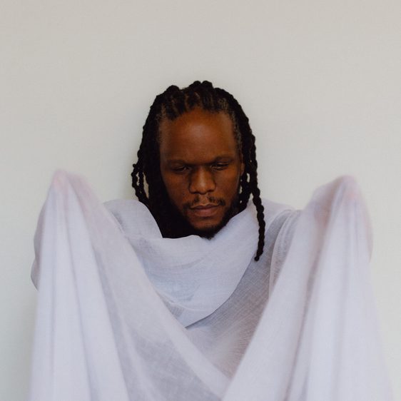 A man with braided hair is holding a sheer, light-coloured fabric in front of him. He appears contemplative, with his eyes closed, and is dressed in a simple, flowing garment. The background is plain and neutral, enhancing the focus on him.