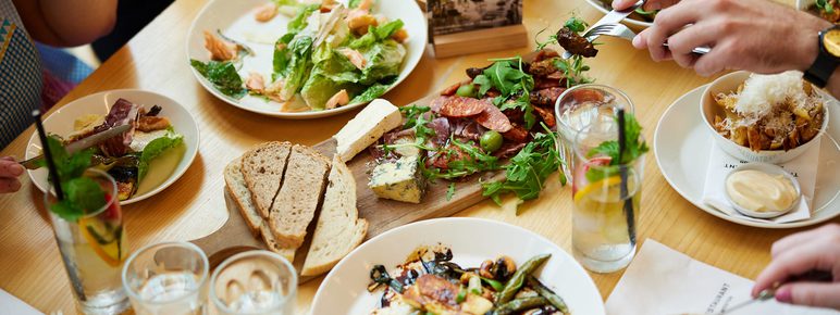 A table filled with small plates of food while hands holding forks reach for the food.