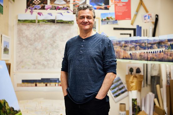 A man with short grey hair standing with his hands in his pockets in an art studio. Behind him are drawings in progress pinned to the walls.