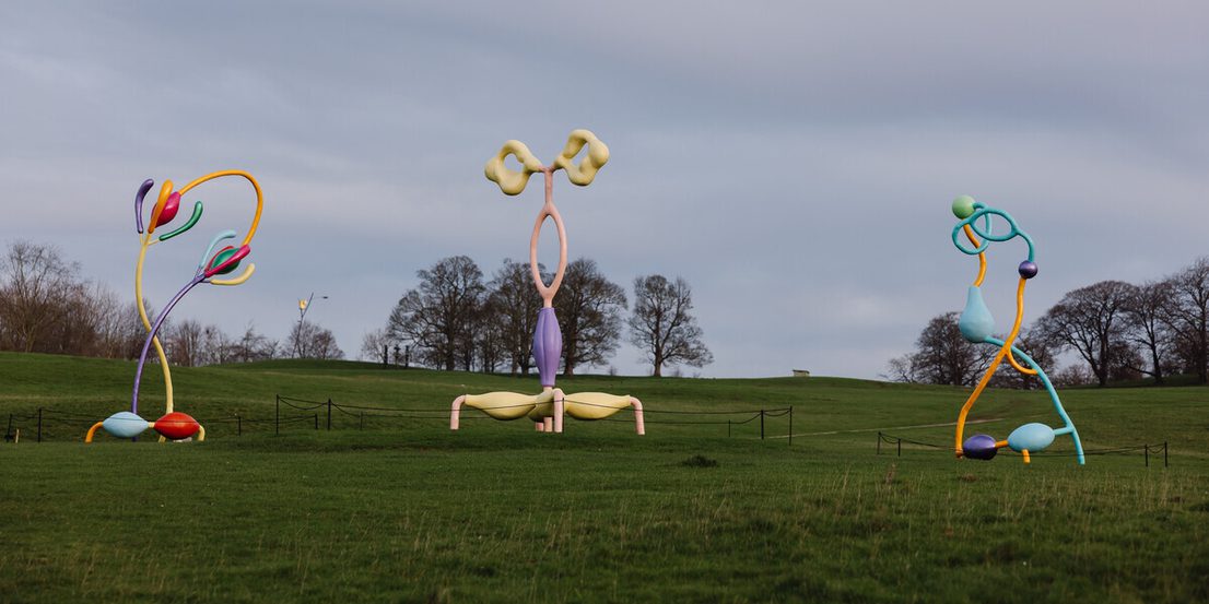 Three colourful abstract sculptures displayed outdoors in parkland, with trees in the distance.