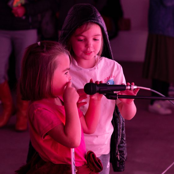 Two children making sounds into a microphone, surrounded by pink light