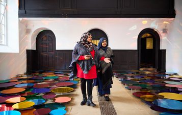 Two women wearing hijab walking along a clear pathway between an installation of colourful mirrored discs.