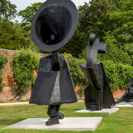 Two large abstract sculptures stand on a grassy area. One features a figure with an oversized, conical head, while the other has a more angular form. Both are made of dark material and are set against a backdrop of lush greenery and trees.