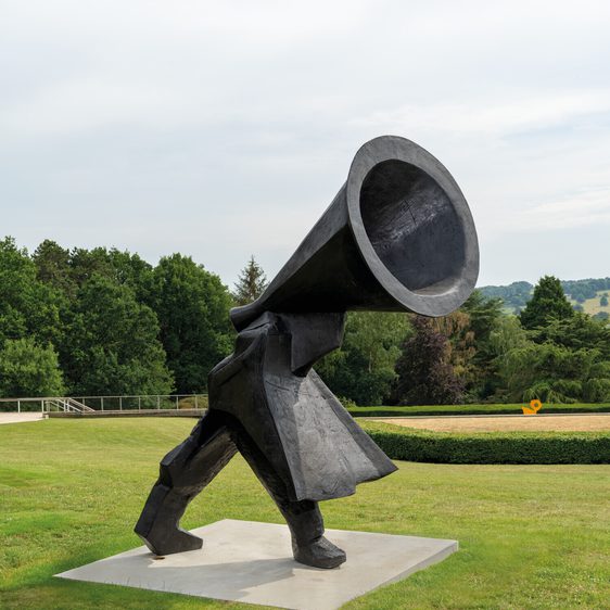 A large, abstract sculpture of a figure walking, with a megaphone-shaped head. The figure is made of dark material and stands on a white base, set against a backdrop of rolling green hills and trees under a cloudy sky.