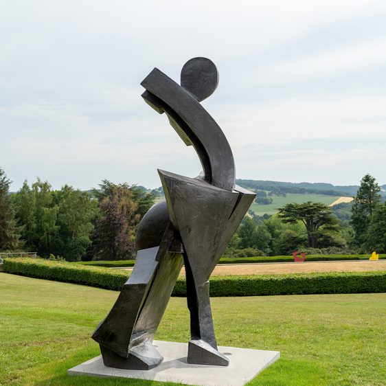 A large abstract sculpture stands in a grassy landscape, featuring geometric shapes and smooth curves. The piece is predominantly black, with a dynamic pose suggesting movement. In the background, rolling hills and trees create a serene setting.