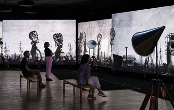 In a dimly lit gallery, three visitors observe large screens displaying monochrome animated figures. Two people sit on wooden benches, while a third stands, all captivated by the striking visuals. Studio lights illuminate the scene.