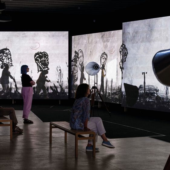 In a dimly lit gallery, three visitors observe large screens displaying monochrome animated figures. Two people sit on wooden benches, while a third stands, all captivated by the striking visuals. Studio lights illuminate the scene.