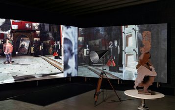 An art installation featuring two large screens displaying a gallery scene with various artworks. A sculptural figure stands on a small table, while a vintage spotlight illuminates the space, creating a dramatic atmosphere.
