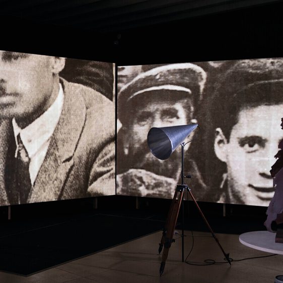A dimly lit exhibition space features two large screens displaying historical black-and-white photographs of men, one in a suit and the other in military attire. In the foreground, a vintage spotlight on a tripod illuminates a sculptural piece.