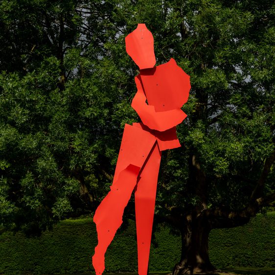 A large, abstract sculpture in bright red, depicting a stylised human figure in a dynamic pose. The figure is composed of angular, geometric shapes and stands on a black base, surrounded by lush green trees and grass.