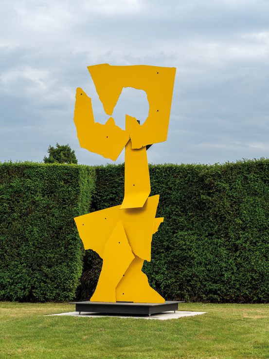 A tall, abstract sculpture made of bright yellow metal stands in a green garden. The piece features angular shapes and cut-outs, contrasting with the neatly trimmed hedges in the background under a cloudy sky.