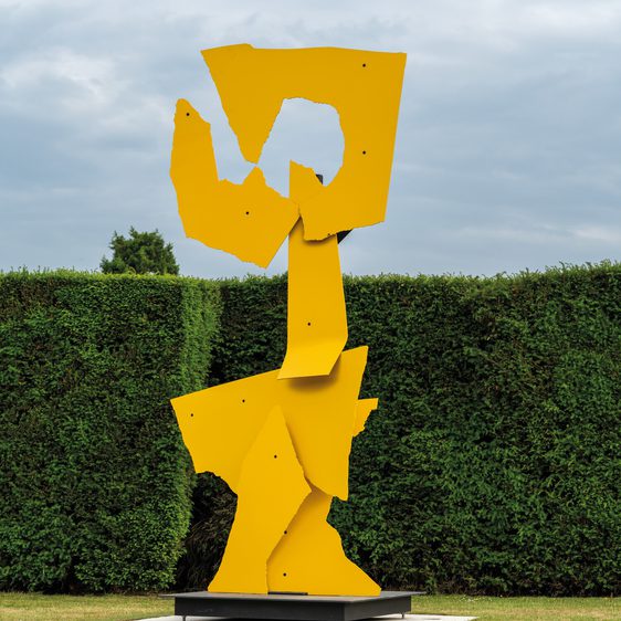 A tall, abstract sculpture made of bright yellow metal stands in a green garden. The piece features angular shapes and cut-outs, contrasting with the neatly trimmed hedges in the background under a cloudy sky.