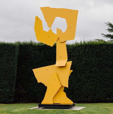 A large abstract sculpture in bright yellow stands against a backdrop of neatly trimmed green hedges. The sculpture features angular, fragmented shapes stacked vertically, creating a dynamic and modern artistic presence in the outdoor space.
