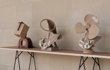 Three bronze sculptures which appear to be made from cardboard are displayed on a wooden table. The first resembles a box, the second features a circular shape, and the third is designed like a fan. Each piece is draped with fabric, adding texture to the display.
