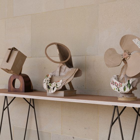 Three bronze sculptures which appear to be made from cardboard are displayed on a wooden table. The first resembles a box, the second features a circular shape, and the third is designed like a fan. Each piece is draped with fabric, adding texture to the display.