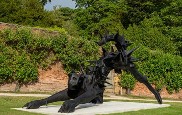 A large, black sculpture of a stylised animal with sharp spikes, positioned on a white plinth. The creature appears to be crawling forward, set against a backdrop of lush greenery and a brick wall, creating a striking contrast.