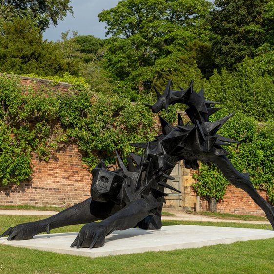 A large, black sculpture of a stylised animal with sharp spikes, positioned on a white plinth. The creature appears to be crawling forward, set against a backdrop of lush greenery and a brick wall, creating a striking contrast.