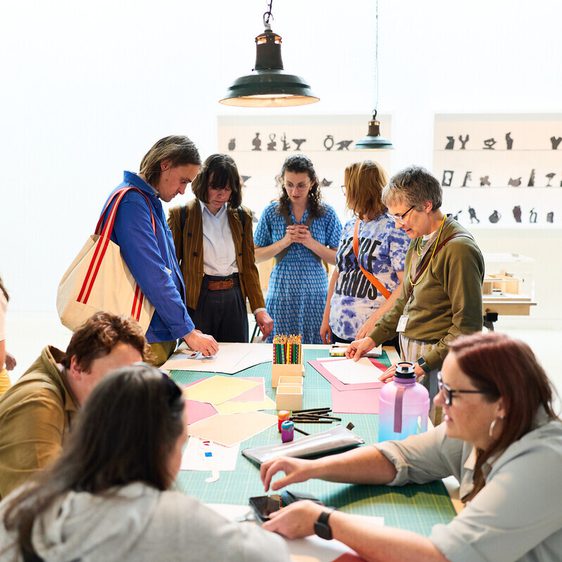 A group of people engage in a creative workshop in a bright, modern space. Some are gathered around a table covered with craft materials, while others are focused on their phones or chatting. The walls feature artistic silhouettes.