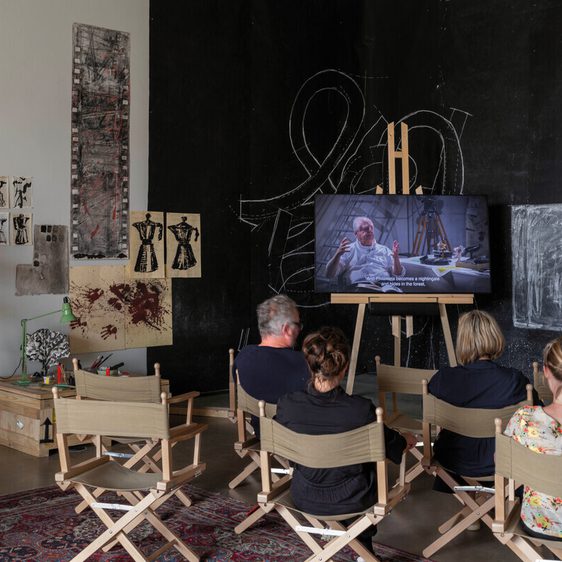 A group of four people sits on canvas chairs, watching a video on a large screen in a creatively decorated room. Behind them, a vintage film camera and various artworks adorn the walls, creating an artistic atmosphere.