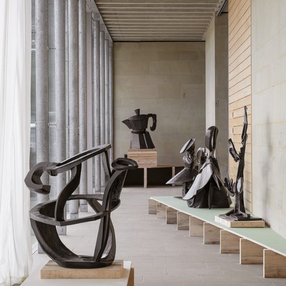 A spacious gallery features several abstract black sculptures displayed on wooden platforms. Natural light filters through large windows, illuminating the minimalist interior with stone walls and a green bench, creating a serene artistic atmosphere.