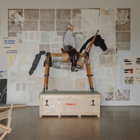 A wooden sculpture of a person riding a horse stands on a crate in an art studio. Behind it, a wall is adorned with various artworks and text, including maps and diagrams, creating a vibrant and eclectic backdrop.
