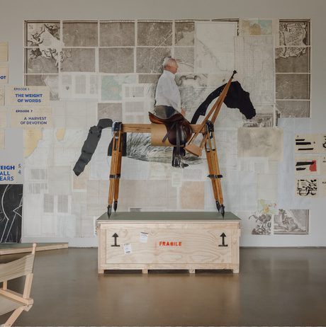 A wooden sculpture of a person riding a horse stands on a crate in an art studio. Behind it, a wall is adorned with various artworks and text, including maps and diagrams, creating a vibrant and eclectic backdrop.