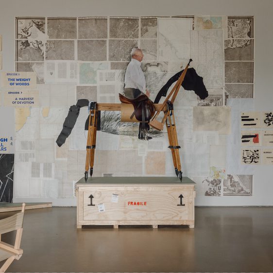 A wooden sculpture of a person riding a horse stands on a crate in an art studio. Behind it, a wall is adorned with various artworks and text, including maps and diagrams, creating a vibrant and eclectic backdrop.