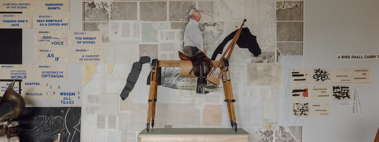 A wooden sculpture of a person riding a horse stands on a crate in an art studio. Behind it, a wall is adorned with various artworks and text, including maps and diagrams, creating a vibrant and eclectic backdrop.