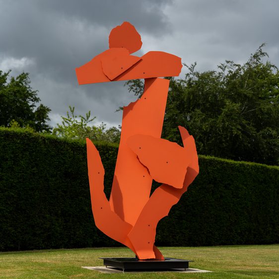 A large, abstract sculpture in bright orange stands prominently on a grassy area. The piece features jagged, angular shapes that suggest human forms, set against a backdrop of lush green hedges and a cloudy sky.
