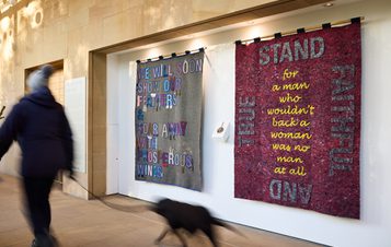 A person walking a dog past a display of fabric banners with embroidered and appliquéd words.