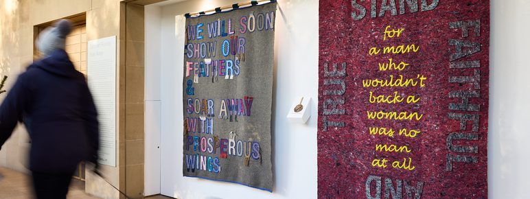 A person walking a dog past a display of fabric banners with embroidered and appliquéd words.