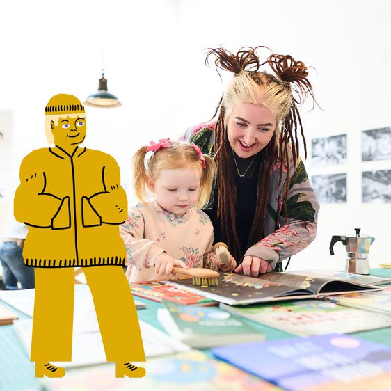 A young girl and a woman with styled hair are engaged in a creative activity at a table covered with colourful books and art supplies. The girl is using a brush on a book, while the woman smiles encouragingly. In the background, a boy is working at another table.