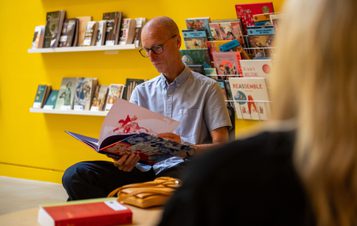 A man reading a book in a yellow room. Rows of children's books are displayed behind him.