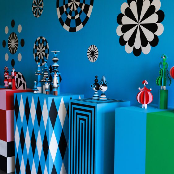 A vibrant exhibition space featuring bold blue walls adorned with black and white geometric patterns. Colourful wooden figures in various shapes and sizes are displayed on multi-coloured pedestals, creating a playful and dynamic atmosphere.