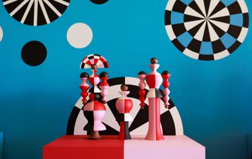 A vibrant display of three abstract wooden sculptures in red, pink, and black, set against a bright blue wall adorned with geometric patterns and circular designs in black, white, and blue. The sculptures feature varied shapes and heights, creating a playful aesthetic.